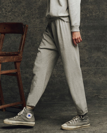 A person in THE GREAT. H25 CORE FLEECEBACK The Fleece Stadium Sweatpant in Heather Grey and gray high-top sneakers walks past a wooden chair against a dark textured background.