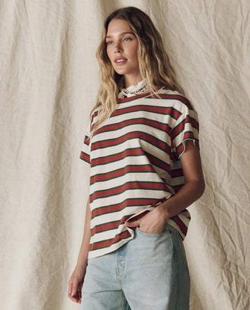 A woman with wavy blonde hair models THE GREAT. F25 D2's The Boxy Crew. Novelty -- Cream Bridle Stripe tee and light blue jeans, standing before a beige fabric backdrop.