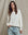 A woman with wavy brown hair wears THE GREAT. CORE KNITS The College Sweatshirt in Washed White with light blue jeans, standing before a beige fabric backdrop.