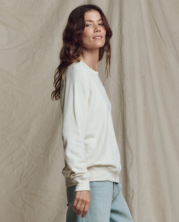 A woman with wavy brown hair stands in profile against a beige fabric backdrop, wearing THE GREAT. CORE KNITS The College Sweatshirt in Washed White and light blue jeans.