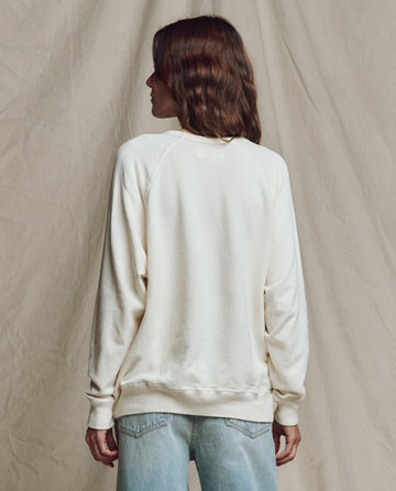 A person with wavy brown hair, wearing THE GREAT. CORE KNITS The College Sweatshirt in Washed White and light blue jeans, stands facing away in front of a beige fabric backdrop.