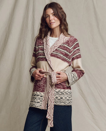 A woman wearing THE GREAT. PF25 The Riding Cardigan in Rusted Red Multi, featuring southwestern motifs in maroon, beige, and cream, stands before a neutral fabric backdrop.