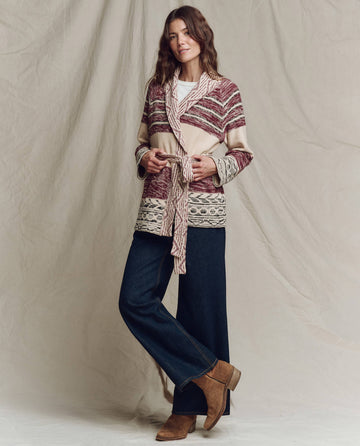 A woman poses in front of a beige backdrop wearing THE GREAT. PF25 The Riding Cardigan in Rusted Red Multi, belted at the waist, paired with wide-leg jeans, a white top, and brown ankle boots.