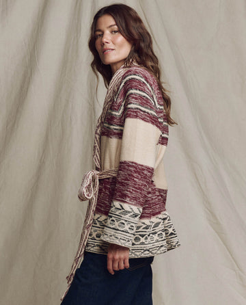 A woman with long brown hair stands sideways in front of a neutral fabric backdrop, wearing THE GREAT. PF25’s The Riding Cardigan in Rusted Red Multi and dark pants.
