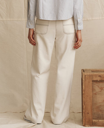 A person stands with their back to the camera, wearing THE GREAT. PS26 The Mill Jean in natural with navy stitching and a blue-and-white striped shirt; a wooden crate sits to the right.