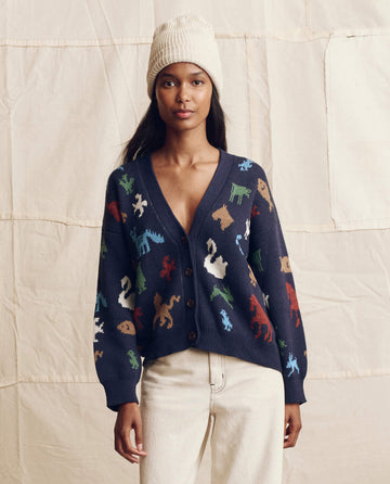 A woman stands before a neutral fabric backdrop, wearing THE GREAT. PS26 The Pictograph Slouch Cardigan in navy, styled with a beige knit hat and cream pants.