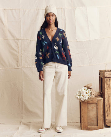 A woman in a cream beanie, cream wide-leg pants, and sneakers wears THE GREAT. PS26 The Pictograph Slouch Cardigan in Navy with colorful animal patterns, standing indoors by flowers and wooden crates.