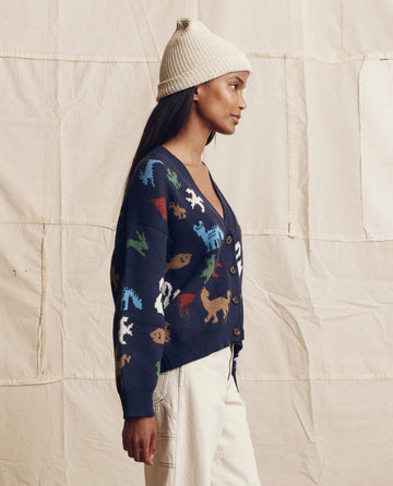 A woman in a cream knit beanie, white pants, and THE GREAT. PS26 The Pictograph Slouch Cardigan in Navy stands in profile before a beige fabric backdrop.