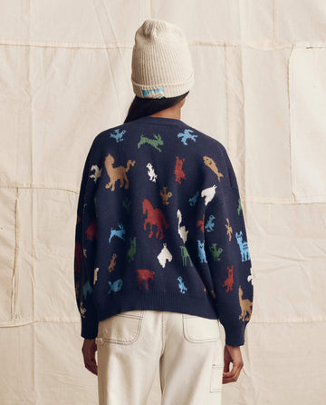 A person faces away, wearing THE GREAT. PS26’s The Pictograph Slouch Cardigan in navy and a cream knit beanie, standing before a neutral fabric backdrop.