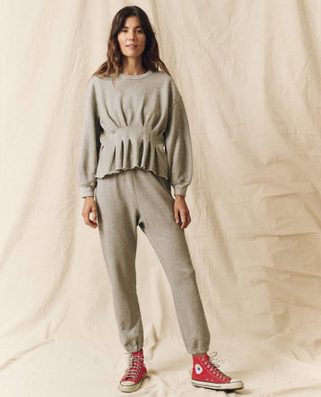 A person stands against a light backdrop, wearing a grey sweatshirt with pleated design and blouson sleeves called "The Corset Sweatshirt - Varsity Grey" from THE GREAT. HOL 23 CORSET SWEATSHIRTS collection, paired with matching grey sweatpants and red high-top sneakers.