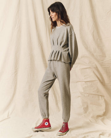 A person standing against a beige backdrop, wearing THE GREAT.’s Varsity Grey Corset Sweatshirt featuring subtle Victorian corset details and matching jogger pants, complemented by red high-top sneakers.