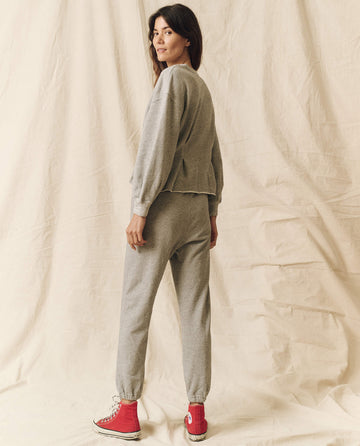 A person stands sideways, looking over their shoulder, dressed in a Varsity Grey Corset Sweatshirt from THE GREAT. HOL 23 collection, paired with sweatpants and red high-top sneakers against a beige fabric background.
