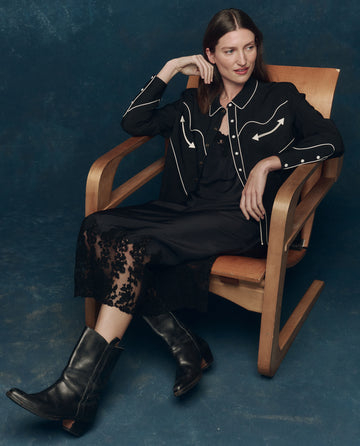 A woman sits in a wooden chair wearing a black dress with lace hem, black boots, and THE GREAT. F25 COLLECTORS "The Western Shirt" in Black with Cream against a dark blue background.