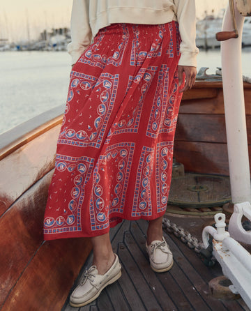 A person stands on a wooden boat deck in THE GREAT. SU26 The Canary Skirt – Red Sailboat Bandana, beige sweatshirt, and light boat shoes; marina and water in the background.