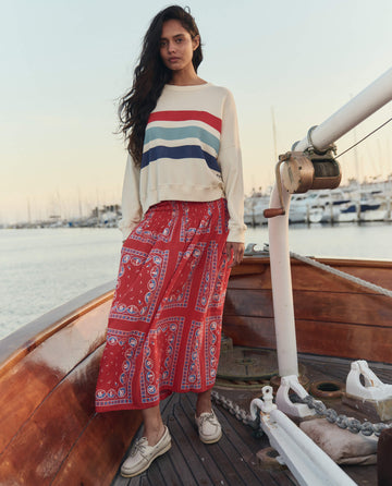 A woman stands on a wooden boat at the marina, wearing THE GREAT. SU26's The Canary Skirt styled with the Red Sailboat Bandana and a cream sweater with colored stripes, docked boats in the background.