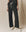 Person wearing loose, washed black The Lap Sweatpant by THE GREAT. CORE KNITS and white high-top sneakers, standing against a beige fabric backdrop.
