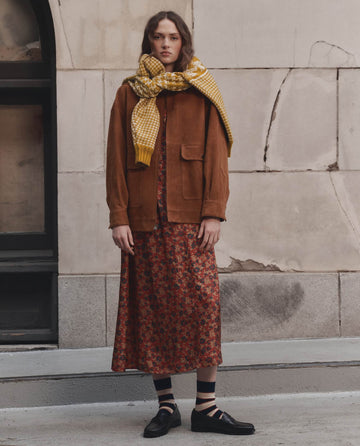 A person stands outdoors against a concrete wall, wearing THE GREAT. F26 D1′s The Suede Folsom Jacket in Cognac, paired with a red floral dress, yellow scarf, black strappy shoes, and striped socks.