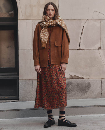 A woman stands against a stone wall wearing THE GREAT. F26 D1 The Suede Folsom Jacket in Cognac, paired with a checked scarf, long floral dress, black socks, and black shoes.