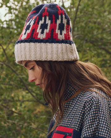 A woman in profile wears THE GREAT. F25 TGO The Nordic Beanie -- Glacier Fair Isle and a patterned jacket, standing outdoors with green foliage in the background.