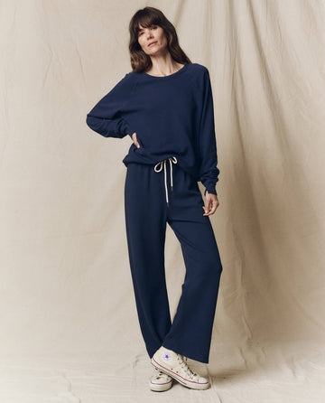 A woman stands against a beige backdrop wearing THE GREAT. CORE KNITS The Lap Sweatpant in True Navy with a matching sweatshirt and white sneakers for a relaxed, stylish look.