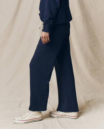 A person stands sideways against a beige fabric backdrop, wearing THE GREAT. CORE KNITS The Lap Sweatpant in Solid -- True Navy with a matching sweatshirt and white high-top sneakers.