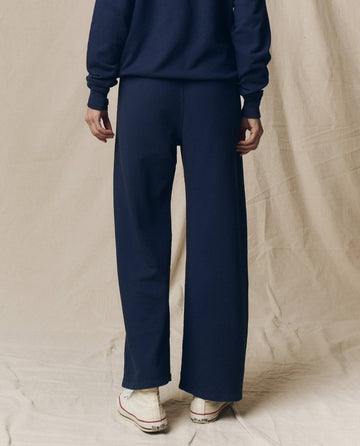 A person stands facing away in THE GREAT. CORE KNITS The Lap Sweatpant. Solid -- True Navy, paired with a navy sweatshirt and white sneakers, against a beige fabric backdrop.