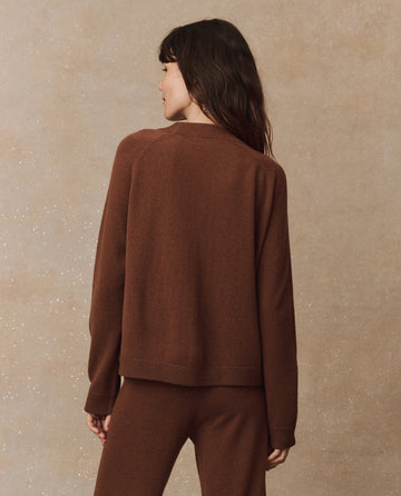 A person with long hair is wearing THE GREAT. HOL 24 CASHMERE's The Cashmere Varsity Cardigan in Santal, paired with pants, and stands against a speckled, light brown background, facing away.