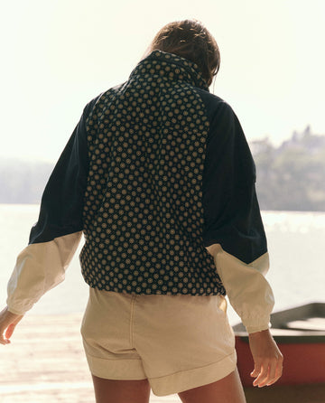 A person wearing The Crestline Full-Zip in Microdaisy Colorblock by THE GREAT. SP25 TGO stands by the waterfront, with trees gracefully framing the serene scene, adding a touch of style.