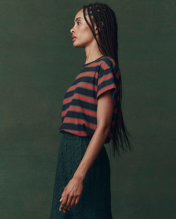 A woman with long braids stands in profile against a dark green background, wearing THE GREAT.'s The Boxy Crew. Novelty -- Navy Bridle Stripe tee and a textured dark skirt.
