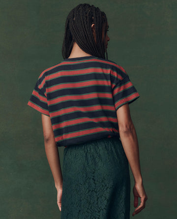 A person with braided hair stands facing away, wearing THE GREAT.'s The Boxy Crew. Novelty in Navy Bridle Stripe against a muted green background.