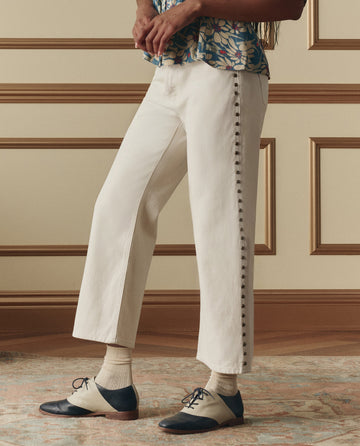 A person stands indoors, lower half visible, wearing THE GREAT. SP26 D2 The Studded Wayne Jean in Natural paired with two-tone oxford shoes, cream socks, and a blue floral top.