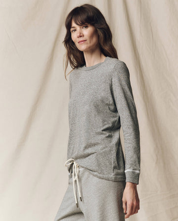 A woman stands before a neutral backdrop in THE GREAT. F25 CORE KNITS The Long Sleeve Slim Tee in solid Heather Grey—an effortless layering staple, paired with matching pants and made in the USA.