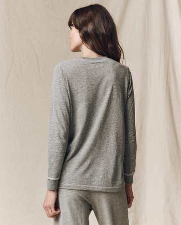 A person with long brown hair stands with their back to the camera, wearing THE GREAT. F25 CORE KNITS The Long Sleeve Slim Tee. Solid -- Heather Grey against a plain beige backdrop—a perfect piece for layering.