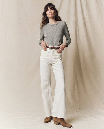 A woman stands before a beige backdrop in THE GREAT. F25 CORE KNITS The Long Sleeve Slim Tee, Solid—Heather Grey, perfectly layered with high-waisted white pants, a brown belt, and tan boots.