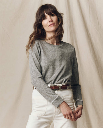 A woman with long brown hair wears THE GREAT. F25 CORE KNITS The Long Sleeve Slim Tee in Solid Heather Grey, tucked into high-waisted white pants with a brown belt, standing against a neutral backdrop. Made in the USA and perfect for layering.