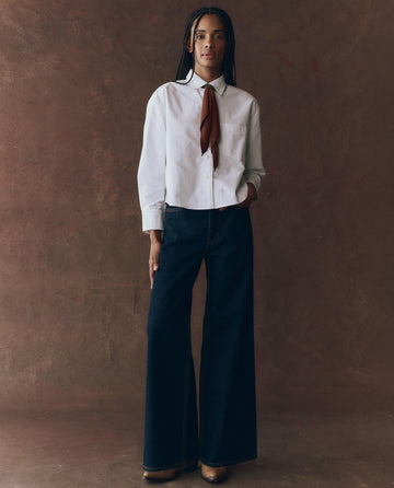 A person stands against a brown textured background, wearing a white button-up shirt with a brown necktie, The Slouch Wide Leg Jean in Midnight Rinse by THE GREAT. F25 D1, and brown shoes.