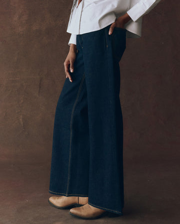 A person wearing a white shirt, THE GREAT. F25 D1 The Slouch Wide Leg Jean in Midnight Rinse, and tan boots stands against a brown textured background.