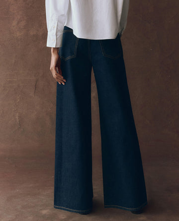 A person stands with their back to the camera, wearing a white shirt and THE GREAT. F25 D1 The Slouch Wide Leg Jean in Midnight Rinse against a brown background.