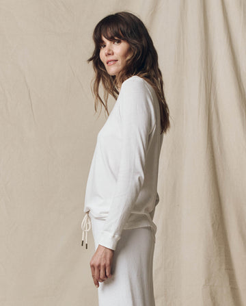 A woman with long brown hair stands sideways in front of a beige fabric backdrop, wearing THE GREAT. F25 CORE KNITS The Long Sleeve Slim Tee in Solid -- True White and a 100% cotton skirt.