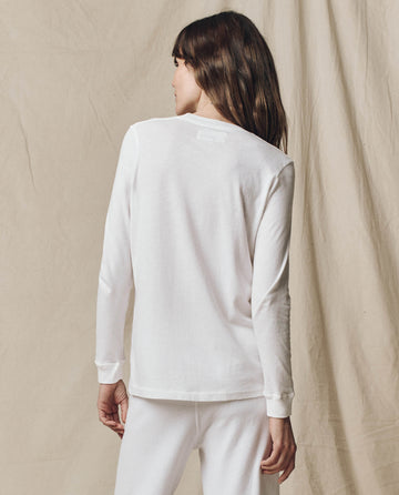 A woman faces away in THE GREAT. F25 CORE KNITS The Long Sleeve Slim Tee, Solid -- True White, paired with white pants against a beige fabric backdrop. Made from 100% cotton, this top delivers classic style and comfort.