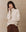 A woman with long hair wears a luxurious ivory turtleneck sweater from THE GREAT. HOL 24 CASHMERE and a brown skirt, standing against a textured background.