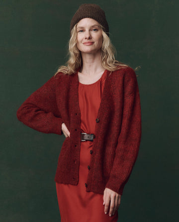 A woman smiles slightly with one hand on her hip, wearing THE GREAT. F25 D2 The Brushed Melange Cardigan in Cinder over a red dress and a brown knit hat, standing against a dark green background.