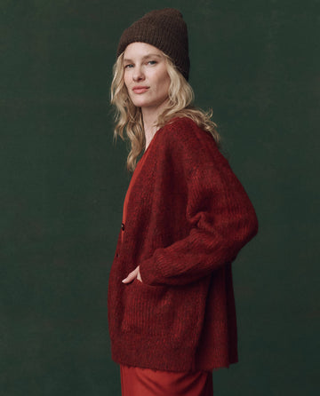 A person wearing THE GREAT. F25 D2 The Brushed Melange Cardigan in Cinder stands against a dark green background, dressed in red with a brown knit beanie, facing slightly to the side with one hand in their pocket.