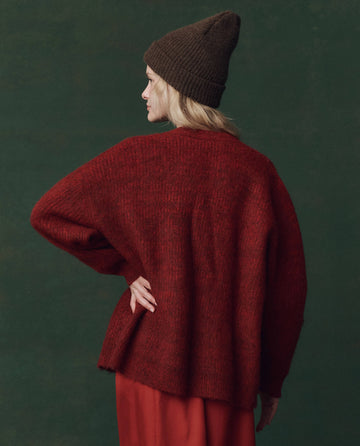 A person wearing THE GREAT. F25 D2 The Brushed Melange Cardigan in Cinder stands with one hand on their hip, facing away from the camera against a dark green background, paired with a brown knit hat.