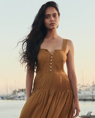 A woman with long dark hair wears THE GREAT. SU26 The Day Break Dress in Clay, standing outdoors by a marina at sunset with boats in the background.