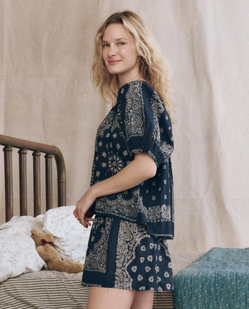 A woman with wavy blonde hair stands indoors, wearing THE GREAT. F25 SLEEP's The Smocked Sleep Short – Bandana Patchwork, a navy blue pajama set with an elastic waist. A teddy bear and patterned blankets on a bed add to the cozy setting.