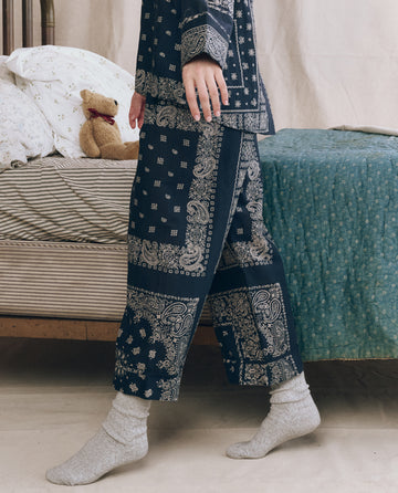 A person wearing THE GREAT. F25 SLEEP The Easy Sleep Pant in Bandana Patchwork stands beside a bed with striped sheets, a greenish blanket, and a teddy bear.