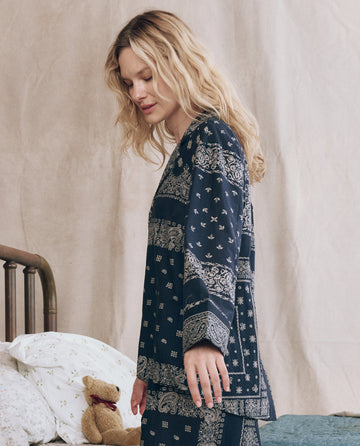 A woman with long blonde hair stands by a bed, wearing THE GREAT. F25 SLEEP's The Pajama Shirt in Bandana Patchwork. Teddy bear and pillows are on the bed in the background.