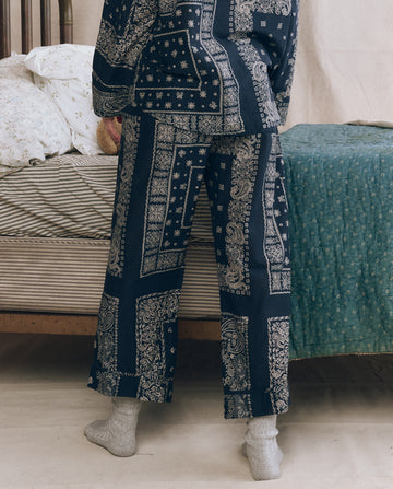 A person stands by a bed in THE GREAT. F25 SLEEP The Easy Sleep Pant - Bandana Patchwork, paired with gray socks, with bedding visible in the background.