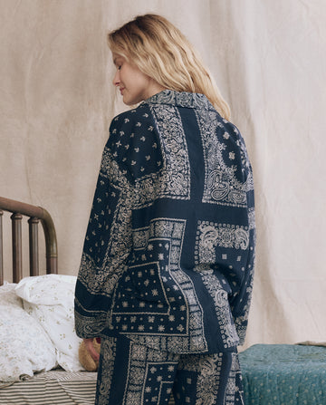 A woman stands beside a bed, facing away, wearing THE GREAT. F25 SLEEP's The Pajama Shirt in Bandana Patchwork.